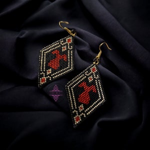 Pode incluir: Um par de brincos de contas em forma de losango. Os brincos apresentam um fundo preto com um design central vermelho e detalhes brancos e dourados. Penduram de ganchos dourados.