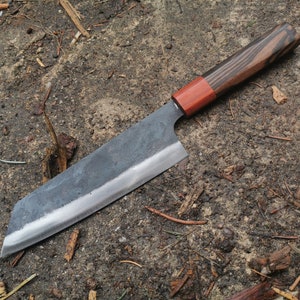 Puede incluir: Un cuchillo artesanal de estilo japonés con una hoja oscura y texturizada y un mango de madera. El mango presenta una virola marrón rojiza y un patrón de vetas de madera a rayas. El cuchillo está diseñado para cortar con precisión.