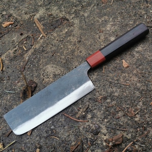 Puede incluir: Un cuchillo de cocina de estilo japonés con un mango oscuro y un acento rojo. La hoja es rectangular y tiene un borde plateado. El cuchillo está sobre una superficie oscura y texturizada.