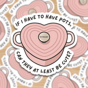 May include: A sticker depicting a pink heart-shaped Le Creuset pot with handles. The text reads, "If I have to have pots, can they at least be cute?" The sticker has a white border and is on a wood-grain background.