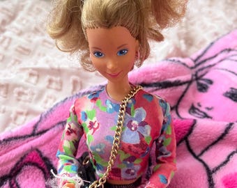 May include: A blonde doll with blue eyes, wearing a floral print top and a pink and white houndstooth skirt. The doll has a gold chain purse and is posed on a pink blanket.