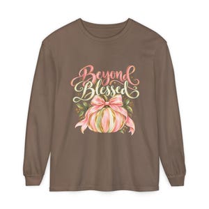 May include: A brown long-sleeve t-shirt featuring a graphic of a pumpkin with a pink bow. The text above the pumpkin reads Beyond Blessed in a stylized pink and gold script font. The design includes green leaves and gold accents.