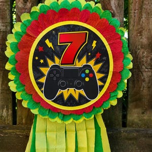 May include: A round pinata featuring a red number 7 and a black video game controller graphic on a yellow starburst background. The pinata is decorated with layers of red, green, and yellow crepe paper fringe and includes long yellow and green streamers.