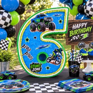 May include: A number 6 shaped blue piñata with green fringe, decorated with monster truck and motocross graphics, featuring the text STUNT ZONE. The design includes checkered flags, stars, and lightning bolt accents on a bright blue background.