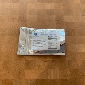 May include: A silver foil package of "Farm Fresh Organic Freeze Dried Raw Eggs" sits on a wooden butcher block. The package includes a nutrition facts label and text that reads "1 dozen eggs". The package is sealed and ready to use.