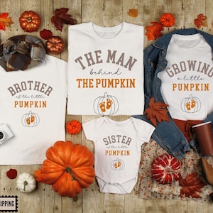 May include: A collection of white t-shirts and a baby bodysuit featuring autumn-themed graphics. The shirts display text including THE MAN behind THE PUMPKIN, BROTHER of the little PUMPKIN, SISTER of the little PUMPKIN, and GROWING a little PUMPKIN.