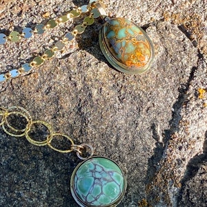 May include: Two necklaces with green and brown stone pendants on a rocky surface. The stones are set in gold and silver.