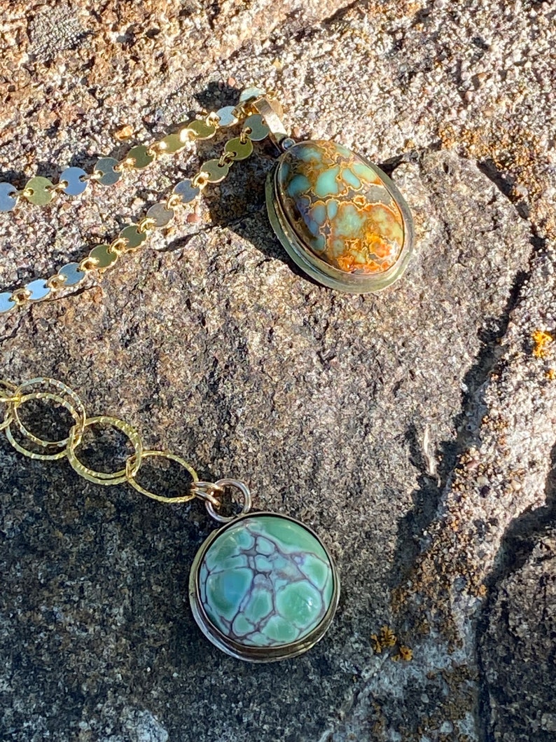 May include: Two necklaces with green and brown stone pendants on a rocky surface. The stones are set in gold and silver.