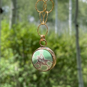 May include: A gold chain necklace with a green and brown stone pendant. The pendant is round and has a textured surface. The necklace is hanging against a blurred background of green trees.