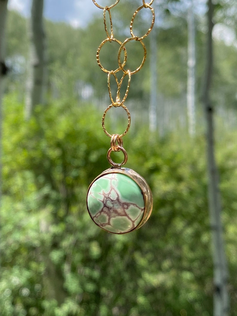 May include: A gold chain necklace with a green and brown stone pendant. The pendant is round and has a textured surface. The necklace is hanging against a blurred background of green trees.
