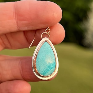 May include: A teardrop-shaped turquoise gemstone earring with a silver setting and a gold accent. The earring is on a gold hook.