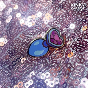 Puede incluir: Un pin de esmalte azul y morado con forma de botella con un tapón en forma de corazón. El pin tiene un contorno dorado y está sobre un fondo rosa brillante. El texto "Kinky Karrot" es visible en la esquina superior derecha de la imagen.