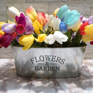 May include: A metal planter with the words "Flowers & Garden" printed on the side. The planter is filled with a bouquet of colorful artificial tulips in shades of yellow, pink, purple, red, white, and blue.