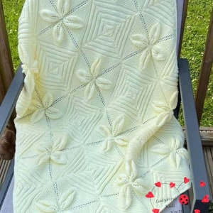 May include: A crocheted baby blanket in a pale yellow colour with a repeating pattern of flowers. The blanket is draped over a chair on a wooden deck.