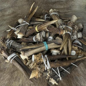 May include: A collection of deer antlers, varying in size and shape, are bundled together with white string and rubber bands. The antlers display a range of brown and tan hues, with some sections appearing lighter in color. The background is a textured, fur-like material.