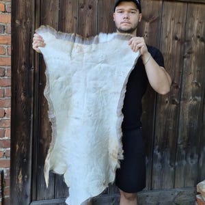 May include: A tanned animal hide with a creamy white center and a border of light brown fur. The hide is irregularly shaped and appears to be soft and pliable. The hide is being held up in front of a wooden door.