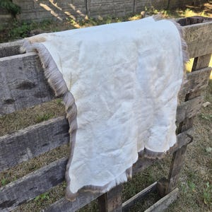 May include: A cream-colored hide with a gray fur trim is draped over a wooden structure. The hide has a soft, textured appearance. The fur trim adds a natural, rustic element to the piece. The hide is likely used for decorative purposes.