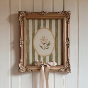 May include: A framed watercolor painting featuring a floral design. The artwork is set within a gold ornate frame, and the background has green and white vertical stripes. A beige satin bow is tied at the bottom of the frame.