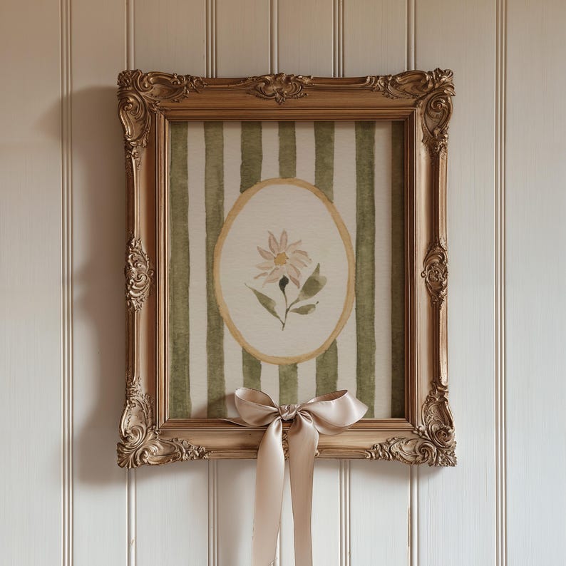 May include: A framed watercolor painting featuring a floral design. The artwork is set within a gold ornate frame, and the background has green and white vertical stripes. A beige satin bow is tied at the bottom of the frame.