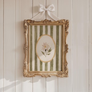 May include: A framed artwork featuring a watercolor painting of a daisy inside an oval, set against a green and white striped background. The ornate gold frame is suspended by a white ribbon tied in a bow, creating a vintage aesthetic.