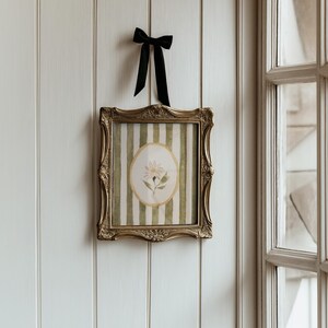 May include: A framed floral print with a gold ornate frame is hanging on a white wall. The print features a daisy in an oval frame against a green and white striped background. A black ribbon is tied above the frame.