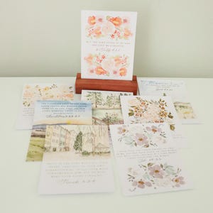 May include: A collection of watercolor greeting cards with floral designs and inspirational quotes. The cards feature soft pastel colors, including pink, peach, and green. The cards are arranged on a white surface, with one card displayed in a wooden holder.