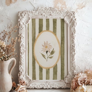 May include: A framed watercolor print featuring a daisy within an oval, set against a green and white striped background. The ornate white frame complements the soft colors of the artwork. Decorative flowers and a vase are in the foreground.
