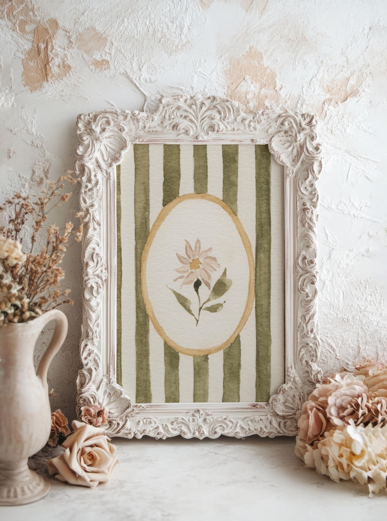 May include: A framed watercolor print featuring a daisy within an oval, set against a green and white striped background. The ornate white frame complements the soft colors of the artwork. Decorative flowers and a vase are in the foreground.