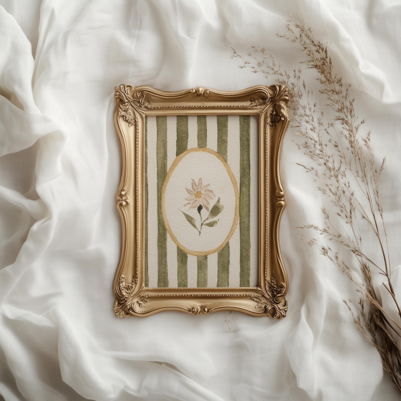 May include: A gold-colored ornate picture frame with a watercolor painting of a flower. The painting is set against a background of green and white vertical stripes. The frame is resting on a white, textured surface.