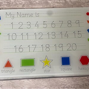 May include: A white dry-erase board with a dotted line grid for writing numbers and the words "My Name is". The board also has colourful shapes including a red hexagon, a blue square, a yellow star, a green rectangle, a red triangle, a green square, a yellow star, a blue square, and a red pentagon.