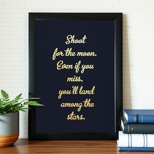 May include: Black framed print with the quote "Shoot for the moon. Even if you miss, you'll land among the stars." in gold script on a navy blue background. The print is displayed on a wooden surface with books and a potted plant.