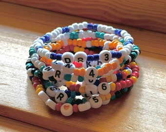 Könnte beinhalten: Ein Stapel bunter Perlenarmbänder mit weißen Perlen, auf denen Buchstaben und Zahlen stehen. Die Armbänder liegen auf einer Holzoberfläche.