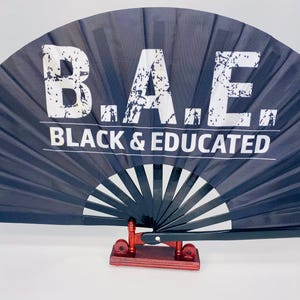 May include: A black folding fan with the text "B.A.E. BLACK & EDUCATED" in white. The fan is displayed on a small, red-brown wooden stand. The fan's design features white radiating lines.