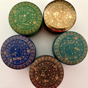 Könnte beinhalten: Fünf runde, dekorative Behälter mit Sternzeichen und Sternbildern. Jeder Behälter zeigt ein anderes Sternzeichen: Krebs, Löwe, Widder, Jungfrau und Waage. Die Behälter sind grün, blau und schwarz mit goldenen Akzenten.