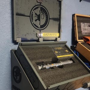 May include: A gray and black wall mount display case with a lightsaber inside. The case has a Star Wars Rebel Alliance symbol on the front and the words "Mera Jade" on the side.