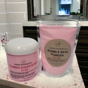 Może przedstawiać: Dwa różowe produkty do kąpieli, peeling cukrowy z pianą i proszek do kąpieli z bąbelkami, oba oznaczone "Love Spell" przez Ebony Botanicals. Peeling znajduje się w przezroczystym słoiku z białą pokrywką, a proszek w przezroczystym plastikowym woreczku.