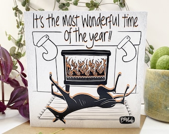Könnte beinhalten: Eine Grußkarte mit dem Text "It's the most wonderful time of the year!!" zeigt eine schwarze Katze, die sich vor einem Kamin entspannt. Die Illustration ist in Schwarz und Orange auf weißem Hintergrund, mit einem braunen Umschlag.