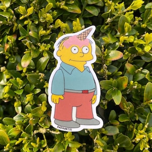 Könnte beinhalten: Ein Aufkleber mit einer Zeichentrickfigur mit gelber Haut, die ein blaues Hemd und eine rote Hose trägt. Die Figur hat rosa Eiscreme mit einer Waffel auf dem Kopf. Der Aufkleber befindet sich auf einem Hintergrund aus grünen Blättern. Der Text "BADMANTIS" steht unten.