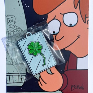 Peut inclure: Un porte-clés rectangulaire avec un motif de trèfle à quatre feuilles vert scintillant. Le porte-clés est tenu devant une illustration de dessin animé d'une personne aux cheveux roux et un fond sombre. Un astronaute de dessin animé est en arrière-plan.