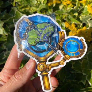 May include: A sticker featuring a stylized, gold-colored scepter with a blue gem and a green and blue ginkgo leaf charm. The scepter has a detailed design with a blue gem at the top. The ginkgo leaf charm is green and blue.