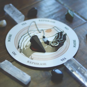 May include: A white and gold divination board with the word "REPHRASE" and the words "YES", "NO", "MAYBE", and "ON". The board is surrounded by crystals and a black pendulum.