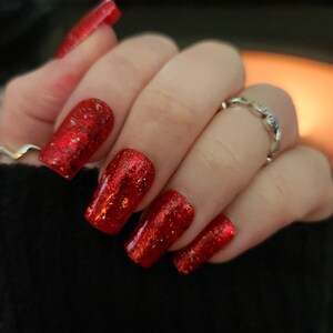 May include: Close-up of a hand with red glitter nail polish on all fingers. The nails are square shaped and the glitter is a fine, red shimmer.