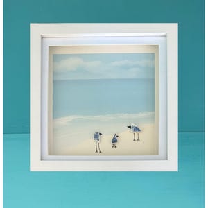 Könnte beinhalten: Ein quadratischer weißer Schattenfugenrahmen mit einer Strandillustration. Drei kleine Vögel aus blau-weiß gemusterten Keramikscherben stehen auf dem sandigen Ufer, mit aufgemalten schwarzen Beinen und Schnäbeln vor einem blauen Ozean.
