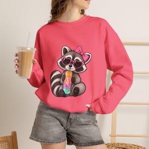 May include: A bright pink long-sleeve sweatshirt featuring a cartoon raccoon wearing pink heart-shaped sunglasses and a pink bow, holding a colourful beverage can with the text Alani printed on it, paired with grey denim shorts.