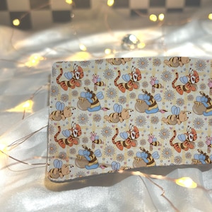 May include: A rectangular fabric item featuring a repeating pattern of cartoon tiger and bee characters, honey pots, and flowers on a light yellow background. The characters are in shades of orange, yellow, and blue. The item is resting on a white, textured surface.
