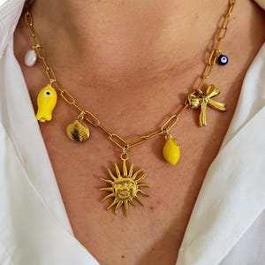 Puede incluir: Un collar de cadena dorada con múltiples dijes, incluyendo un sol, un pez, una concha, un limón, un lazo y un dije de ojo azul.