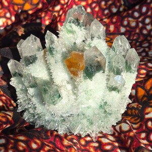 May include: A cluster of clear quartz crystals featuring internal green inclusions and a single prominent amber-colored crystal point. The formation is covered in a dense layer of small, white, needle-like crystal growths across the base.