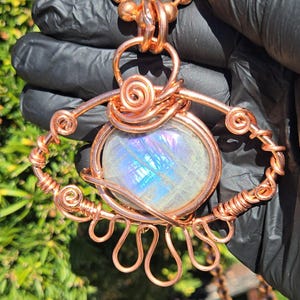 Op de afbeelding: Een handgemaakte koperdraad hanger ketting met een grote, ovale, iriserende edelsteen. De hanger heeft ingewikkelde draadwikkeling met spiraal- en lusontwerpen. De ketting is gemaakt van koperen kralen.