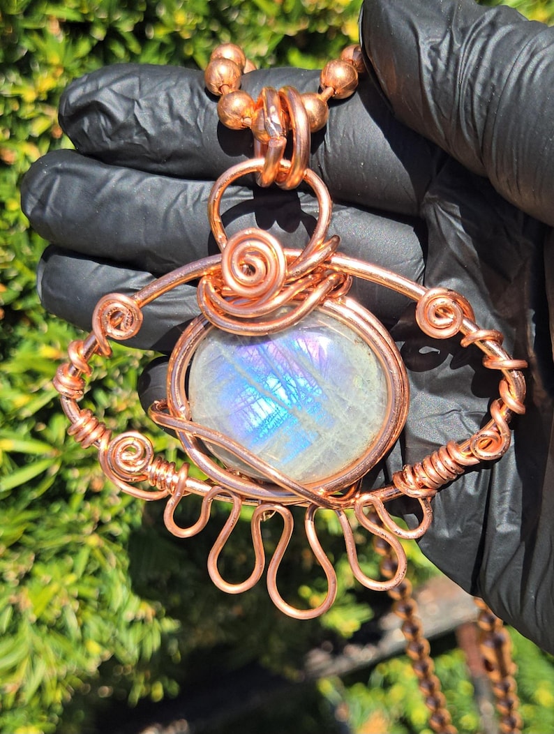 Op de afbeelding: Een handgemaakte koperdraad hanger ketting met een grote, ovale, iriserende edelsteen. De hanger heeft ingewikkelde draadwikkeling met spiraal- en lusontwerpen. De ketting is gemaakt van koperen kralen.
