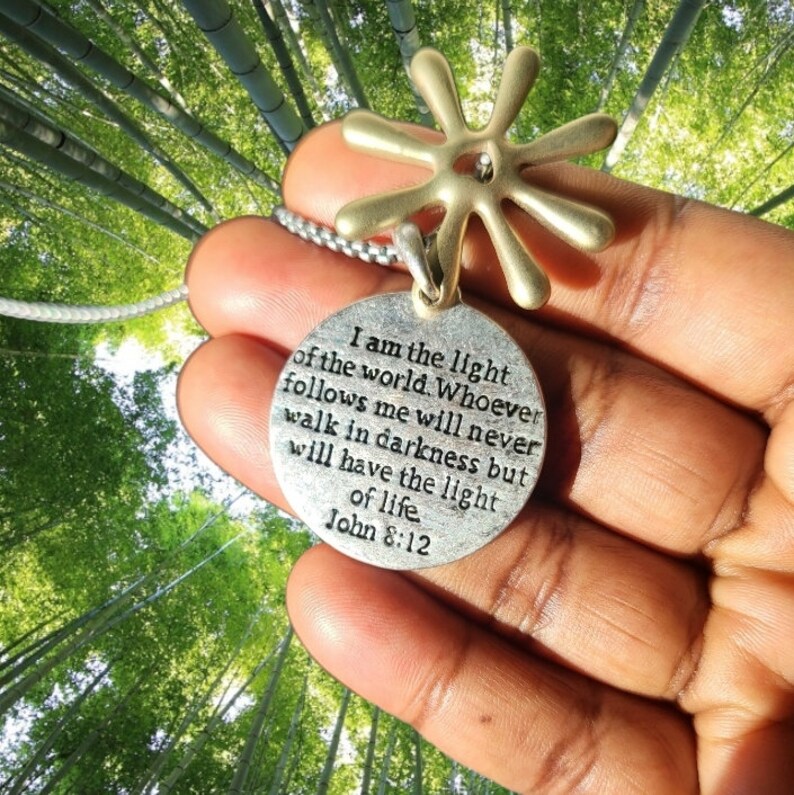 May include: Silver necklace with a round pendant featuring a quote from the Bible, "I am the light of the world. Whoever follows me will never walk in darkness but will have the light of life. John 8:12". The pendant is attached to a silver chain with a gold star-shaped charm.